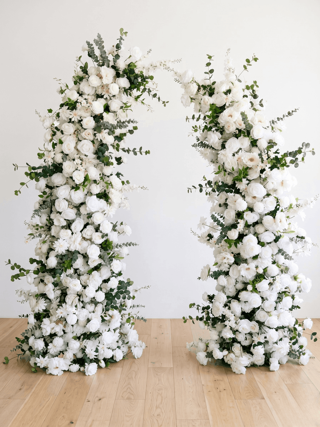 Symmetrical front-on product view of the Bianca broken floral arch columns, showcasing the dense arrangement of premium silk white roses, ranunculus, and trailing eucalyptus on light oak hardwood floors.