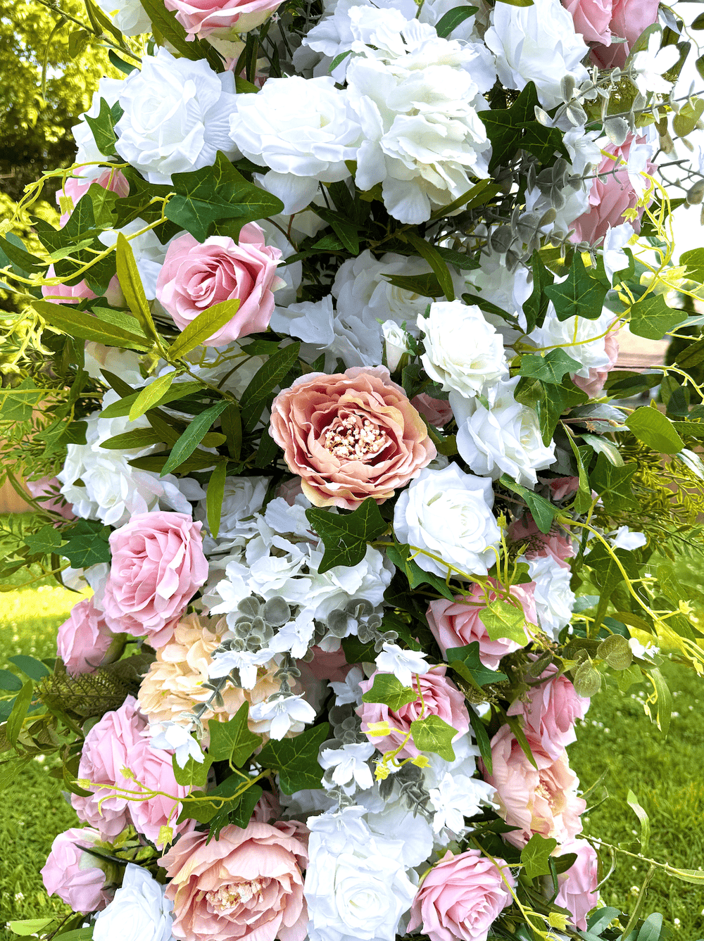 A lush floral arrangement featuring pink and white faux roses, hydrangeas, and green foliage, photographed outdoors against a grassy background.