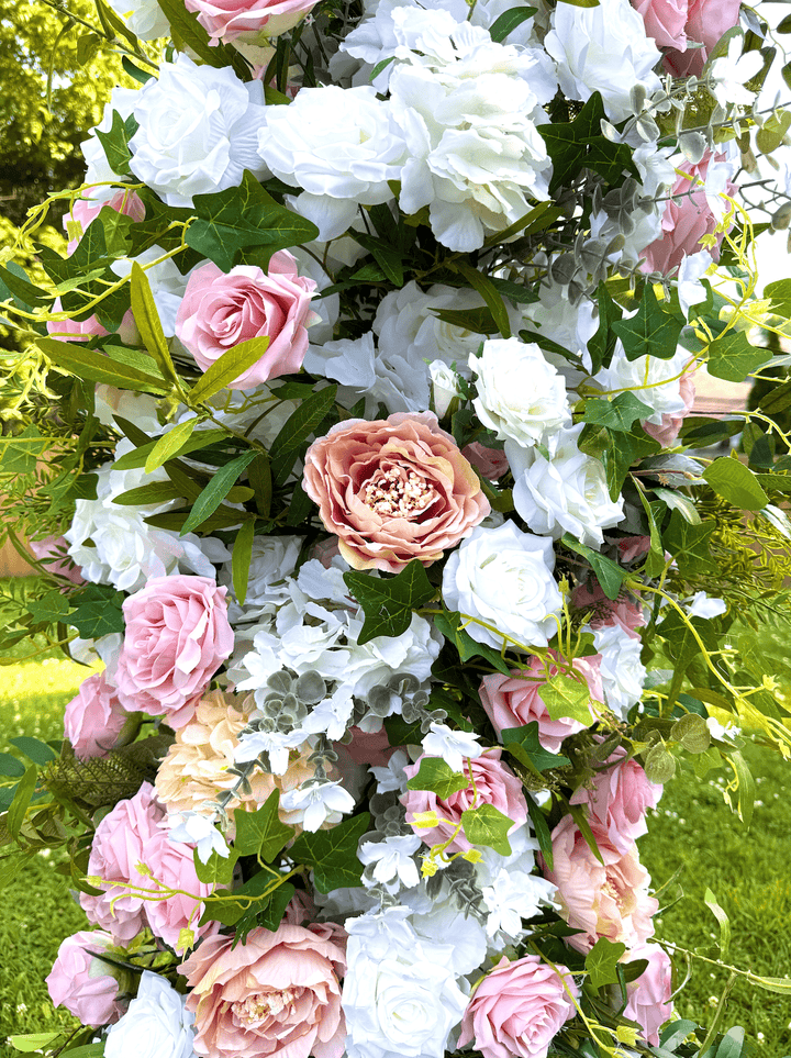 A lush floral arrangement featuring pink and white faux roses, hydrangeas, and green foliage, photographed outdoors against a grassy background.