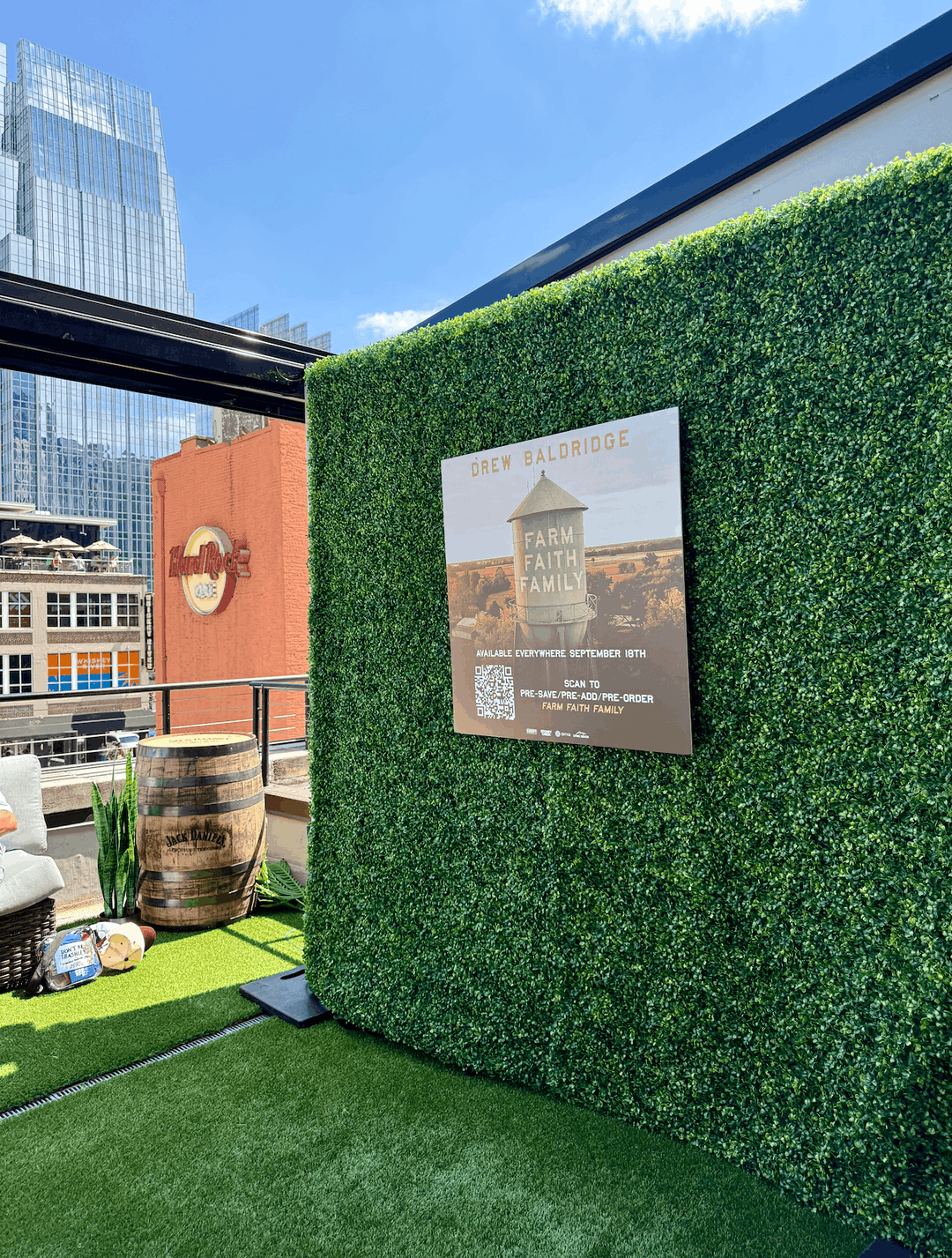 A green artificial hedge wall for sale on a rooftop terrace with a printed sign next to a wooden barrel and city buildings in the background.