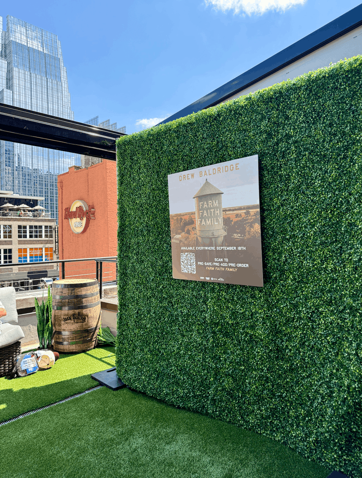 A green artificial hedge wall for sale on a rooftop terrace with a printed sign next to a wooden barrel and city buildings in the background.