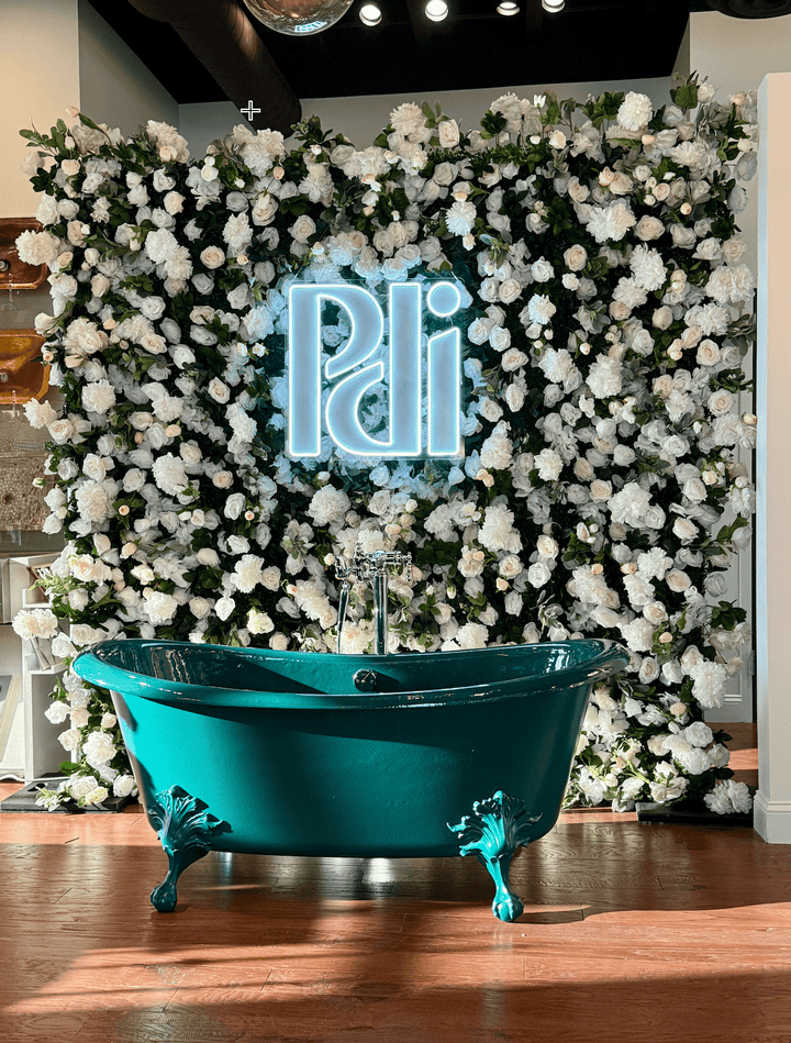 A freestanding teal clawfoot bathtub placed in front of a large white flower and greenery floral wall backdrop with a glowing neon sign, on a wood floor.