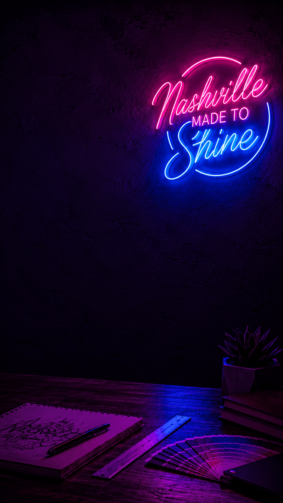 A glowing neon sign on a dark wall reads 'Nashville MADE TO Shine' in pink and blue light, mounted above a wooden desk with books, a notebook, and a mug in a dimly lit room.