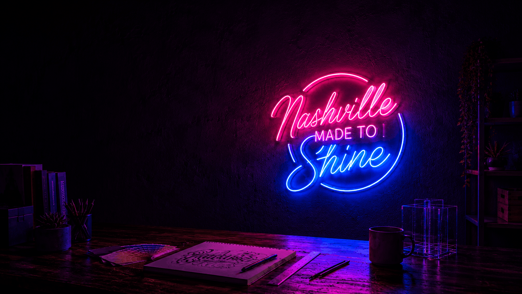 A glowing neon sign on a dark wall reads 'Nashville MADE TO Shine' in pink and blue light, mounted above a wooden desk with books, a notebook, and a mug in a dimly lit room.