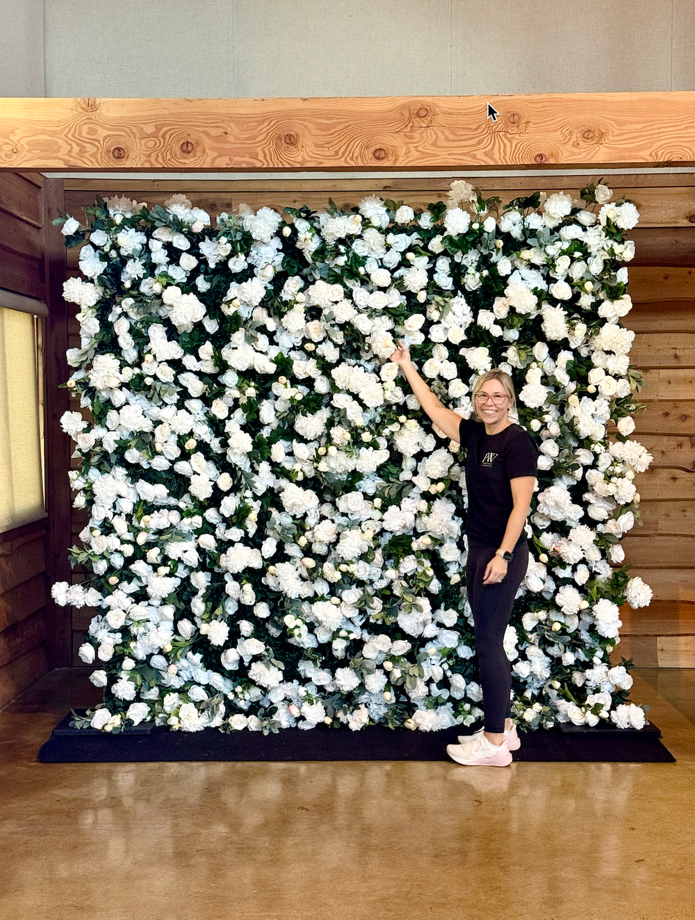 A woman stands in front of a large faux floral wall backdrop made of white flowers and green foliage, posed indoors against a wood-paneled wall and wooden ceiling beam.