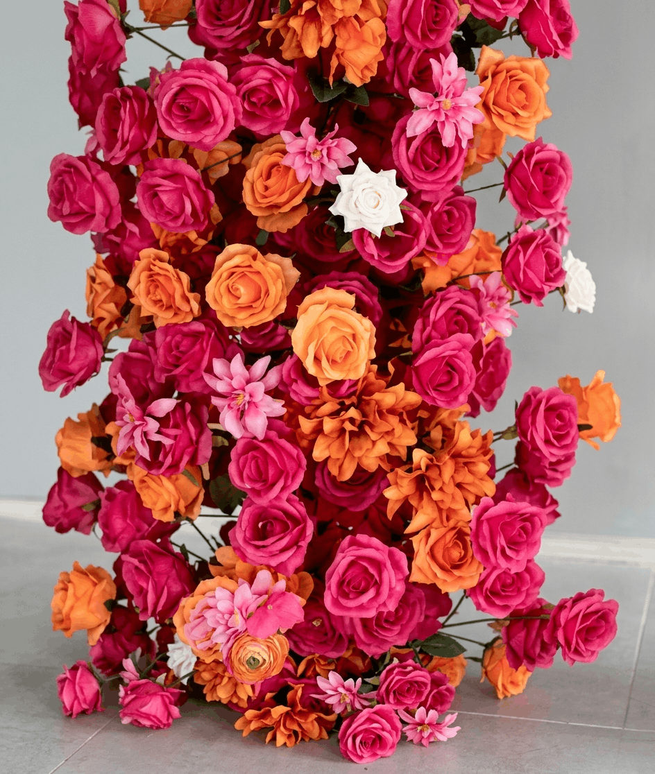 A close up of a flower arch for sale in vivid shades of hot magenta pink, bright orange, and light pink, with a few white blossoms, photographed against a plain light gray wall and floor.