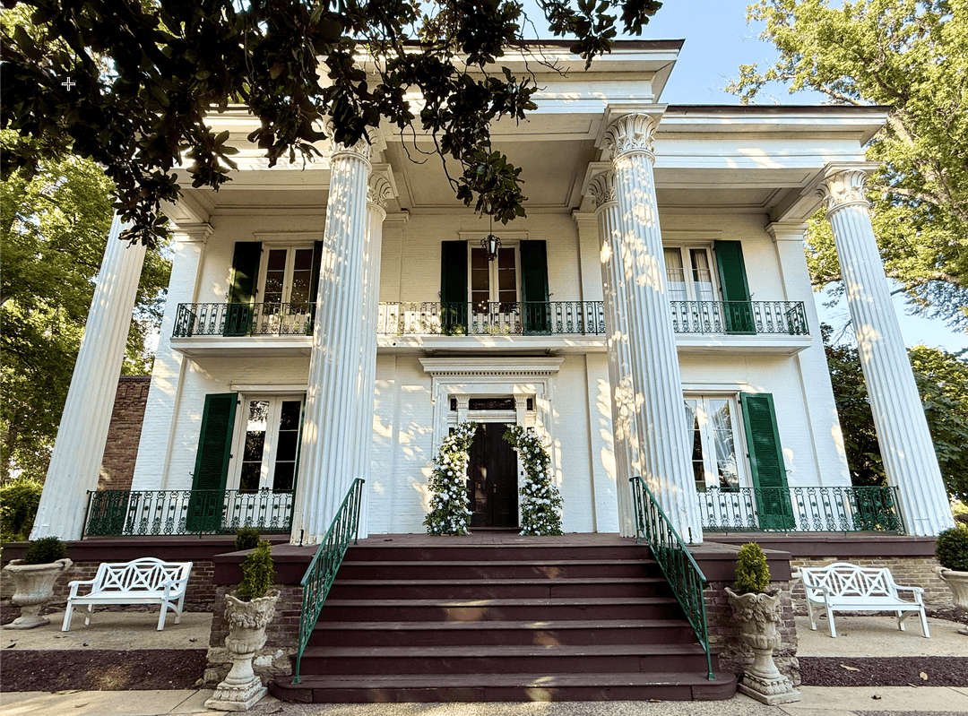 A white and greenery flower arch at grand white plantation-style house with tall columns, dark green shutters, a wide staircase, and trees framing the top of the scene.
