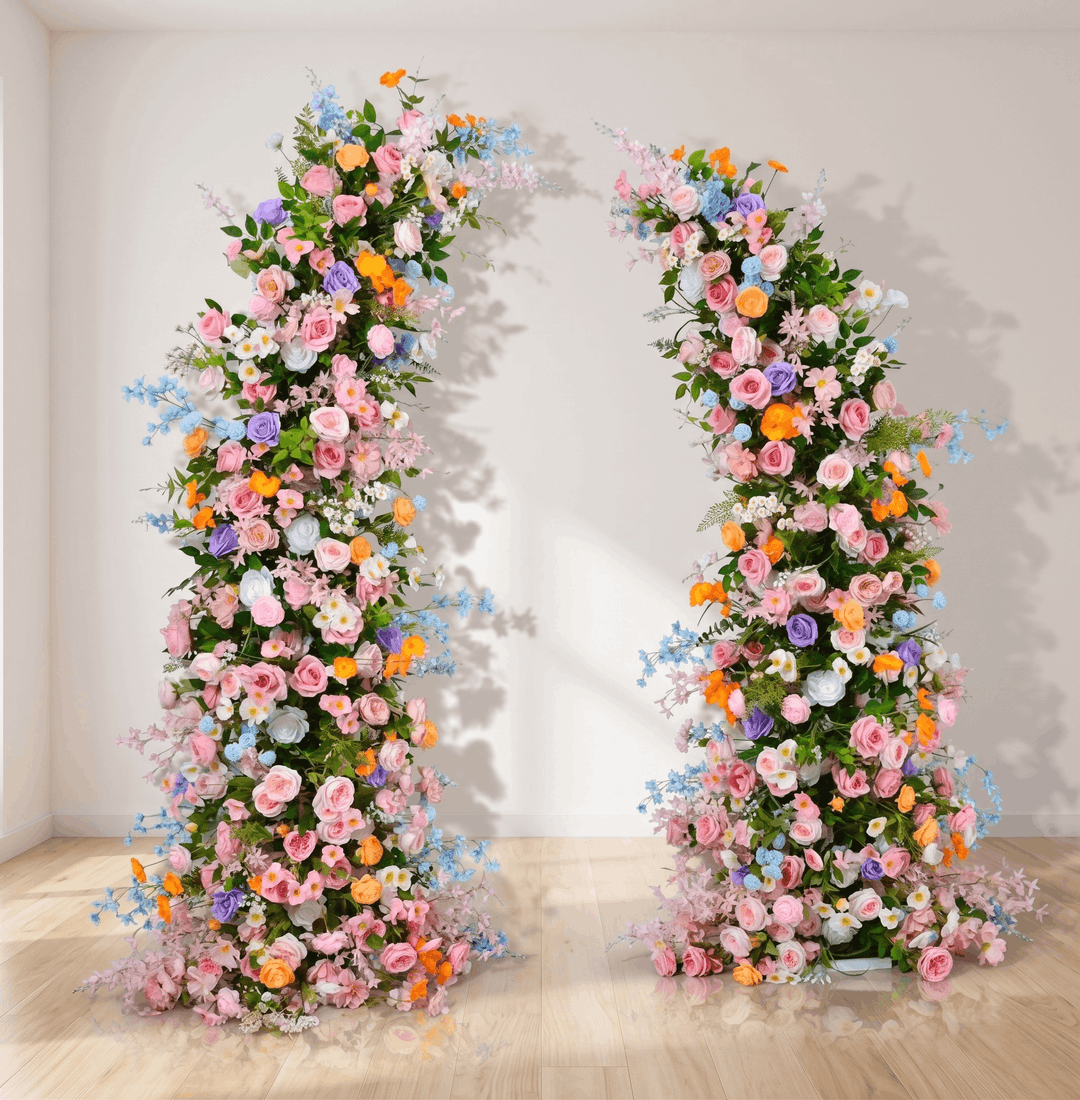 Two tall floral arrangements on a wooden floor with a white wall background