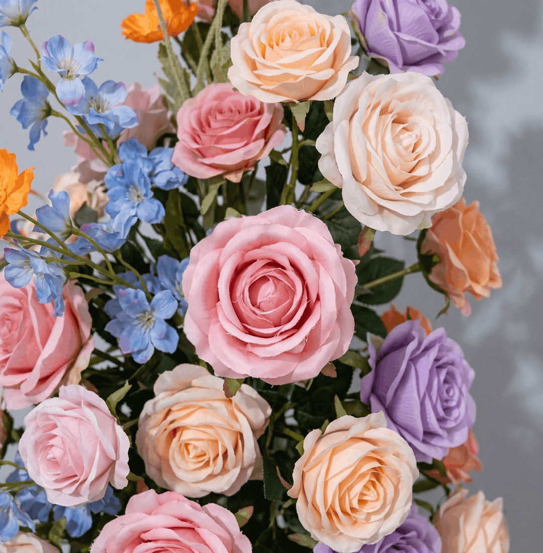 A close-up of a mixed floral arrangement featuring pastel pink, peach, lavender, and blue artificial-looking flowers with green foliage against a soft gray background.