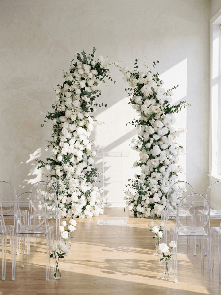 Floral arches with white flowers and greenery in a room with clear chairs.
