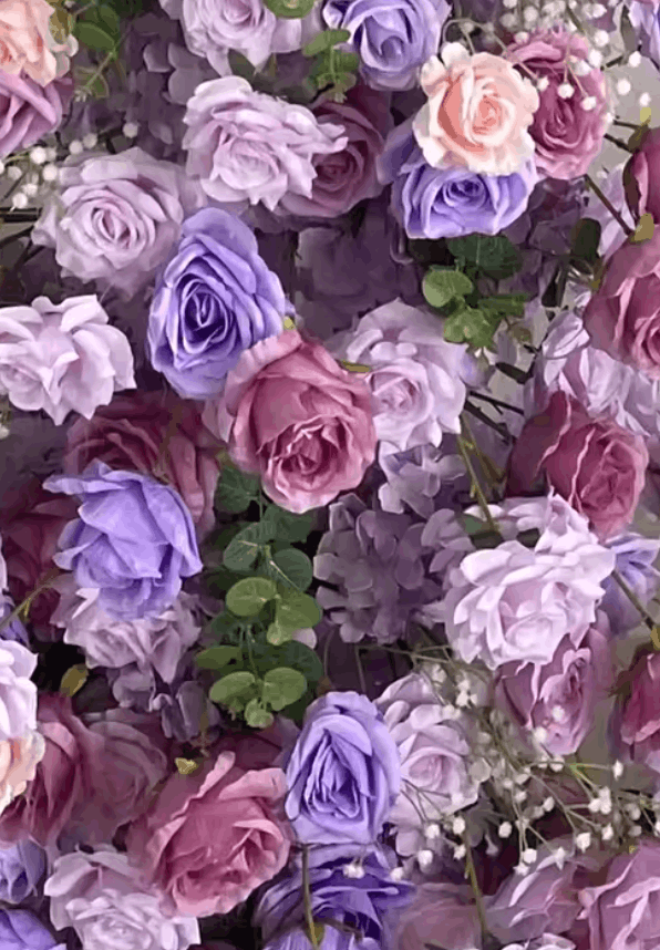 A dense arrangement of artificial roses in soft lavender, lilac, dusty pink, and mauve tones, interspersed with small white baby&