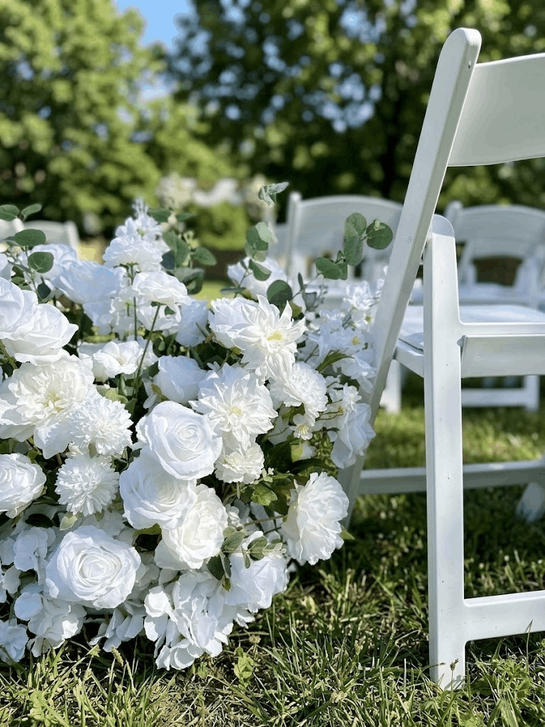 Ground-level floral meadow aisle runner made with white silk roses, hydrangeas, and greenery next to a white wooden ceremony chair.