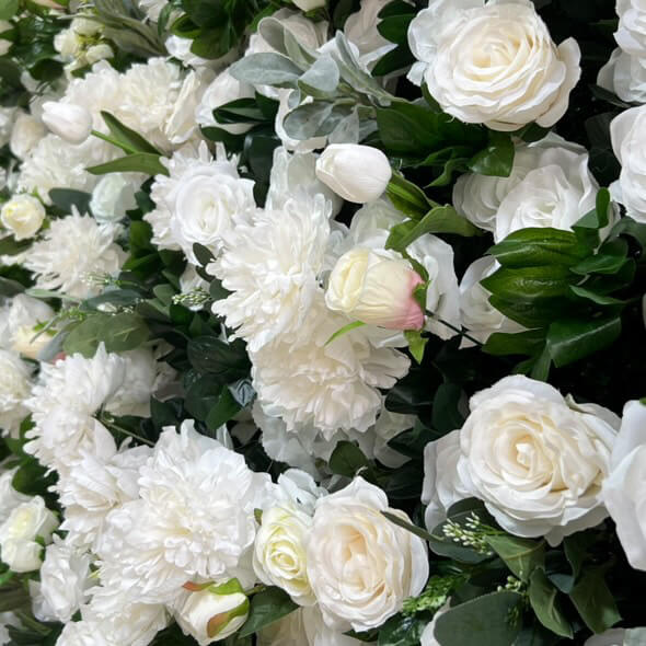 A dense floral wall featuring white and cream roses, peonies, and other white blossoms interspersed with dark green foliage.