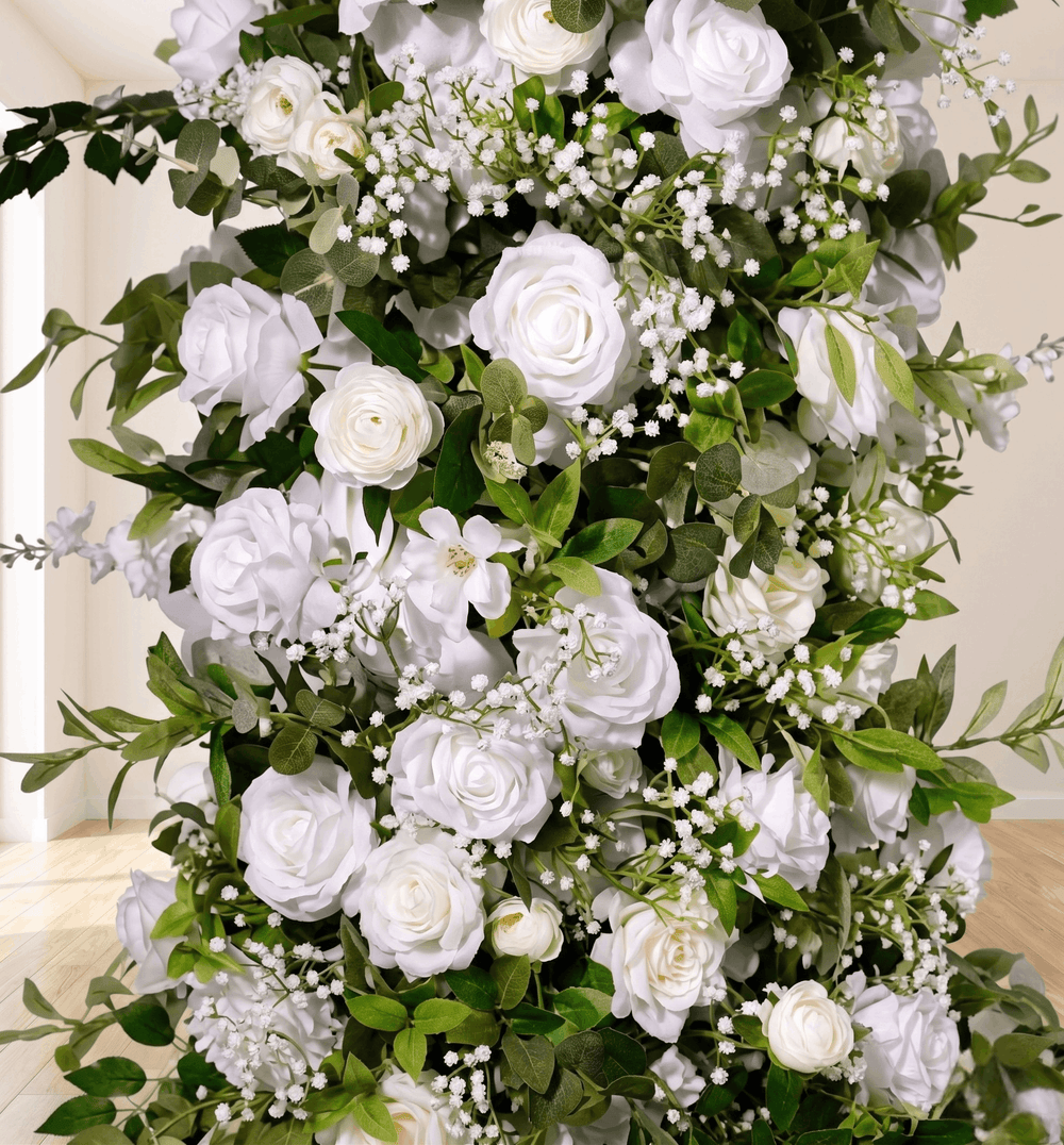 A large artificial floral arrangement featuring white roses, white baby's breath, and green foliage, photographed indoors against a light neutral wall and floor.