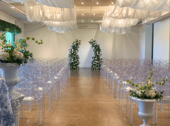 A greenery with white flower arch wedding venue setup with rows of clear acrylic chairs arranged in an aisle, white draped fabric hanging from the ceiling, floral arches at the back, and decorative white pedestal tables with flowers in the foreground.
