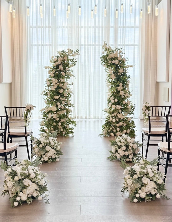 A wedding aisle setup with two tall floral arches and matching flower bouquets in white and green, flanked by black wooden chairs against a bright window backdrop.
