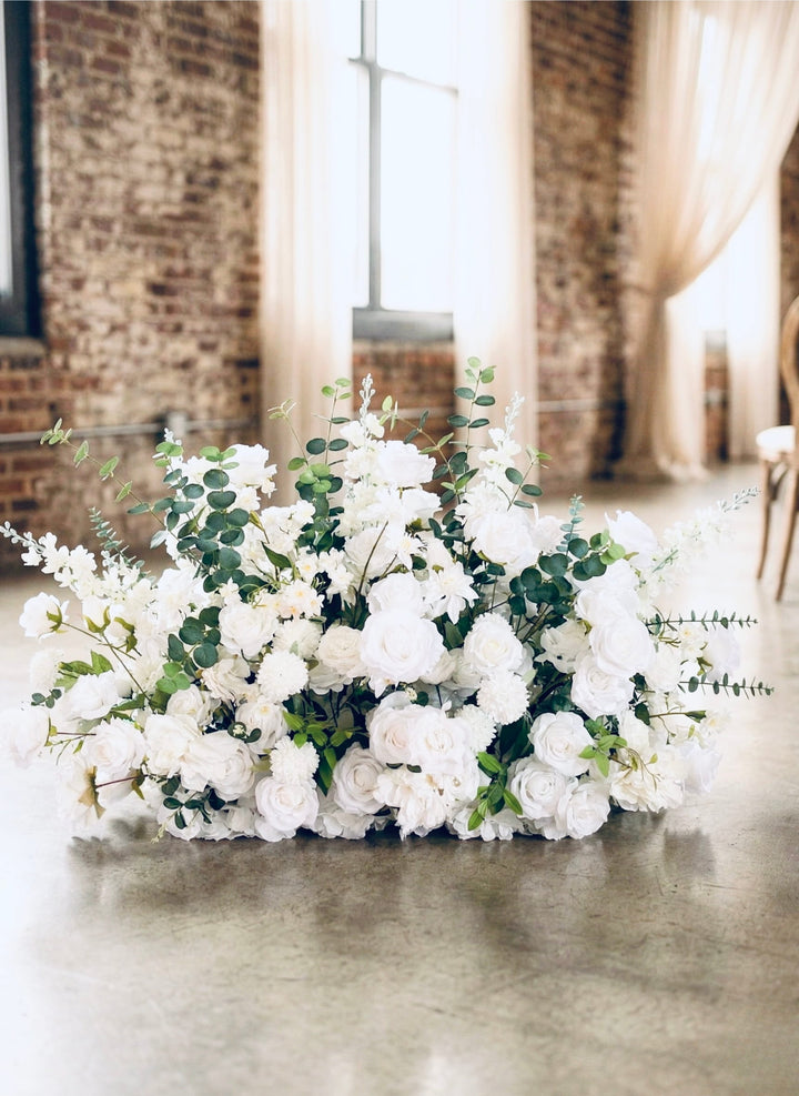 White rose and eucalyptus wedding aisle flower meadow arrangement sitting on a light floor.