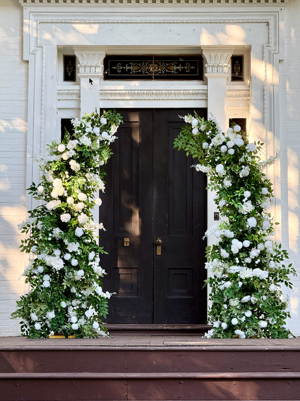 A decorative floral arch made of white flowers and green foliage flanking a dark double door on a white building porch.